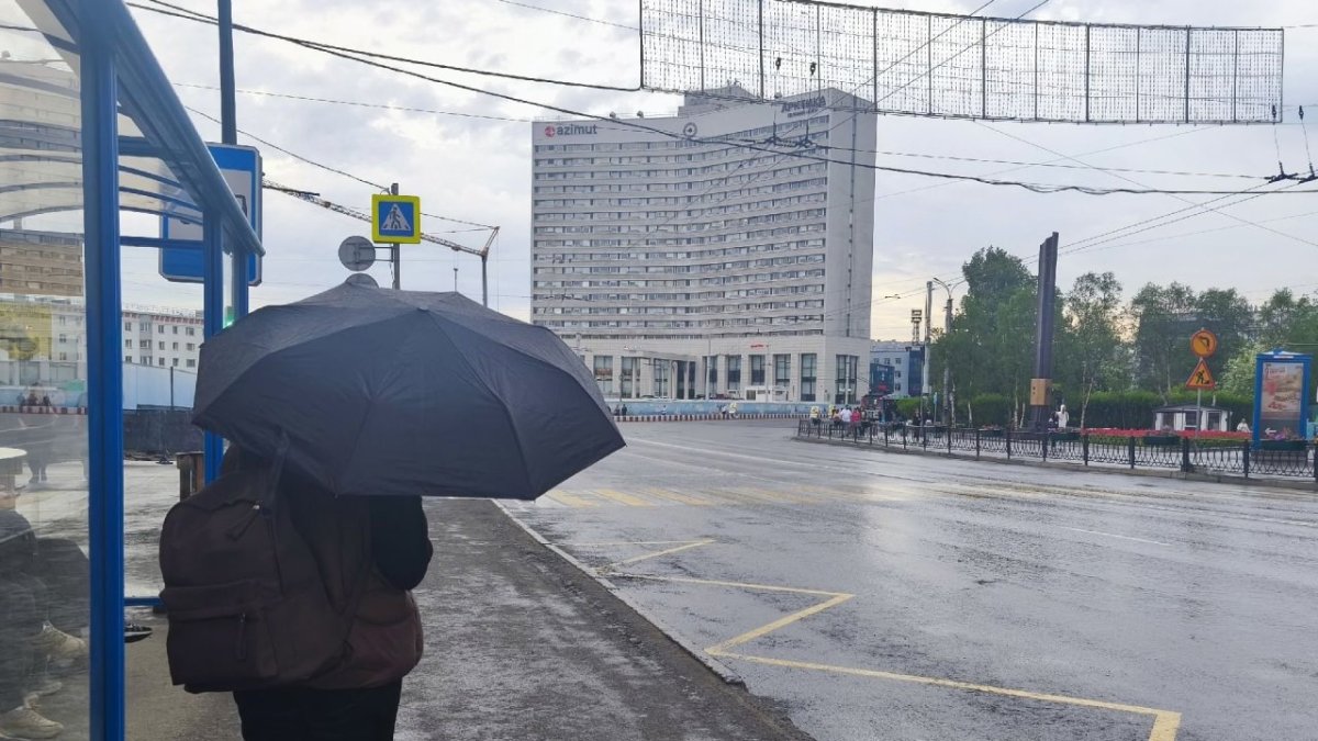 Девушка стоит возле остановки с раскрытым черным зонтом на фоне города