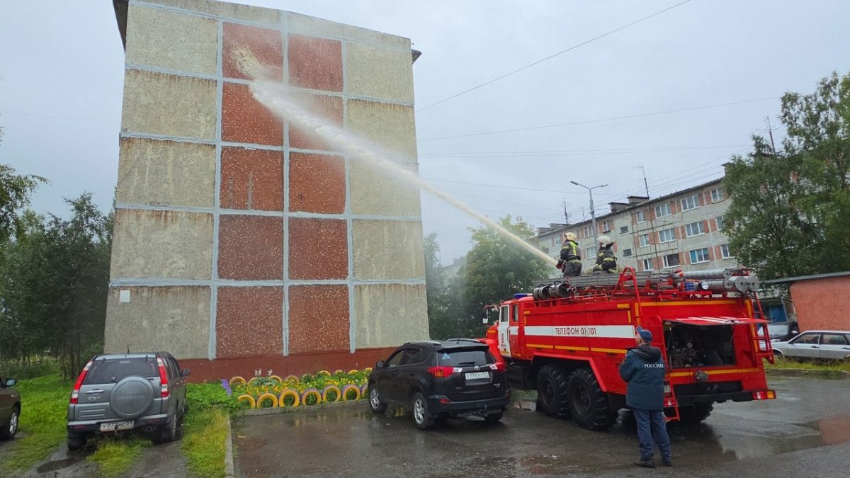 Струя воды из гидранта пожарной машины моет стену жилого дома летом.